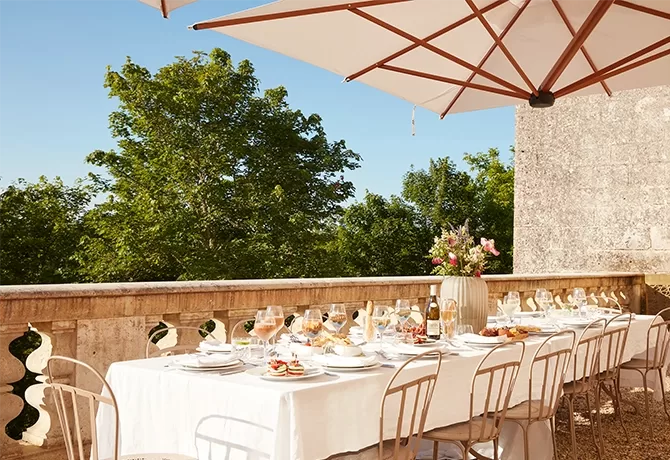 A beautifully set outdoor dining table with a white tablecloth and various dishes is under a large umbrella. Elegant chairs surround the table. In the background, there are lush green trees and a stone wall, suggesting a scenic, sunny setting.