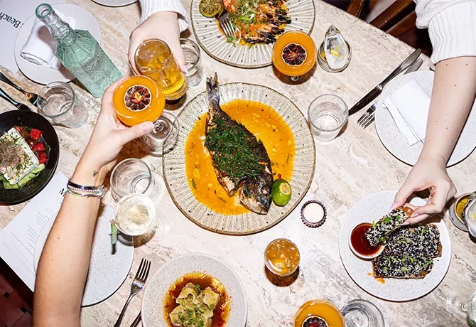 A top view of a dining table with a variety of dishes, including a whole cooked fish, grilled prawns, appetizers, and drinks. Three people are reaching for food and holding glasses, creating a lively and social dining atmosphere at Milk Beach Soho Soho London, London.