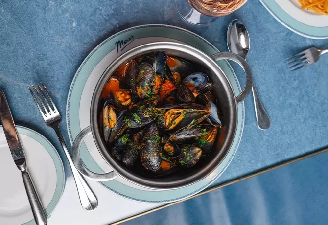 A pot of steamed mussels in a rich broth garnished with herbs, set on a blue table. Silverware is arranged neatly around the dish, and a glass of rosé wine is visible in the background  at Manzi’s, Soho, London.