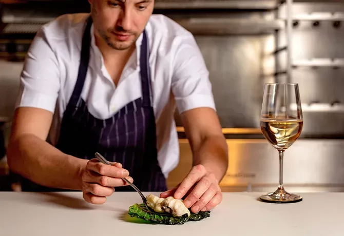 A chef in a white shirt and striped apron carefully plates a dish in a kitchen. A glass of white wine is placed on the counter beside the dish, illuminated under warm lighting at Kebab Queen, Soho, London.