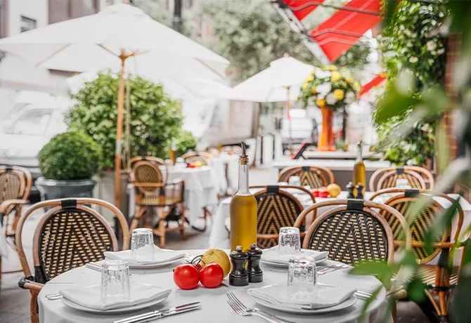 An outdoor restaurant patio with tables set with white tablecloths, plates, cutlery, olive oil, and seasonings. Rattan chairs surround the tables, and red umbrellas provide shade. Tomatoes are placed on one table as decoration at LPM, London.
