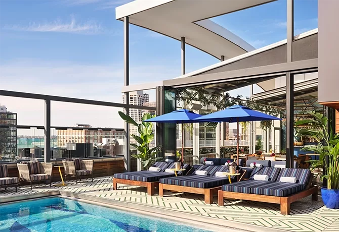 Blue skies overlooking a swimming pool with a glass wall around it overlooking buildings. Blue striped sun loungers with blue parasols on the side.