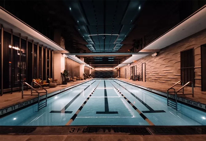 Large lit up swimming pool in a chrome coloured rom with a reflective ceiling showing the reflection of the pool. Floor to ceiling windows show New York in the dark.