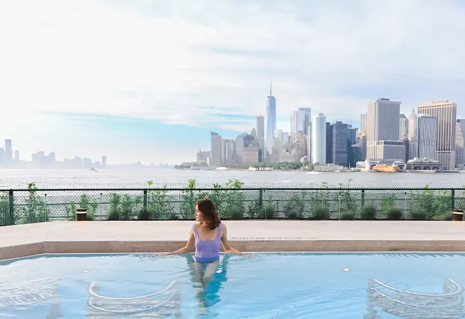 A person in a purple swimsuit relaxes at the edge of an outdoor pool, overlooking a large body of water. In the background, a city skyline with tall skyscrapers is visible under a blue, partly cloudy sky.