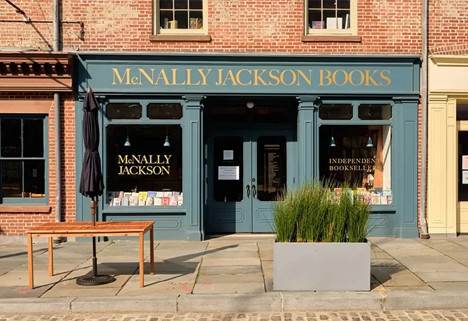 Exterior of McNally Jackson Books in NYC with a green facade and gold lettering. The storefront features large windows displaying books, a small table, a closed umbrella, and a planter with tall grasses on the cobblestone sidewalk.