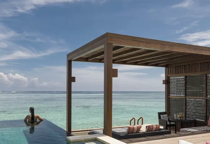 A person relaxes in an infinity pool overlooking a clear blue ocean. A wooden pergola shades a seating area with cushions. A small table holds drinks, set against a backdrop of a sunny sky and calm sea at Four Seasons Maldives at Kuda Huraa, North Malé Atoll.