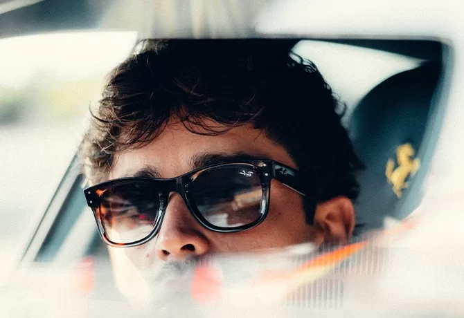 Close-up of Charles Leclerc wearing sunglasses, reflected in a cars rear-view mirror. His expression is relaxed, with soft lighting highlighting his face. A car seat with a logo is visible in the blurred background.