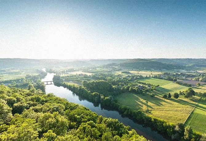 A scenic view of a river meandering through lush green fields and forests under a clear blue sky. A bridge spans the river, with rolling hills and farmland visible in the background in France.