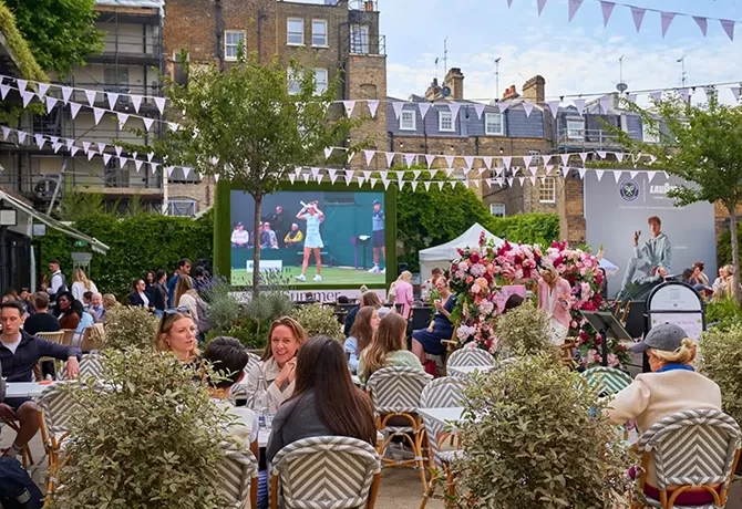 People sit at outdoor tables watching a large screen showing a tennis match, with festive bunting overhead, greenery, and floral decorations in a lively garden setting.