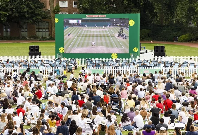 A large crowd sits outdoors on the grass, watching a live tennis match on a giant screen, surrounded by white picket fences and greenery on a sunny day.