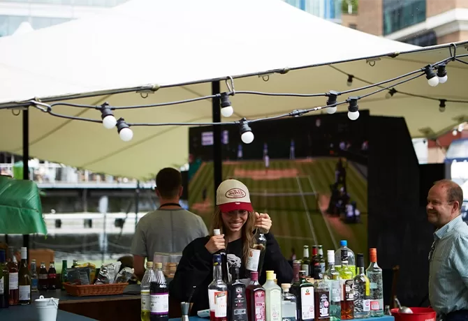 A smiling bartender in a cap gives a thumbs up behind an outdoor bar stocked with bottles, under a white canopy with string lights. Two other people are nearby, and a tennis match shows on a screen in the background.