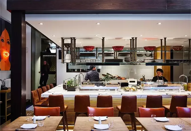A modern restaurant interior with an open kitchen. Chefs are preparing food behind a white marble counter. Red bowls and plates are displayed on shelves above. The dining area features wooden tables and brown leather chairs at Dinings SW3, London.