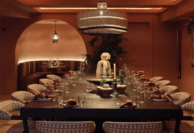 A dimly lit, elegantly set dining room with a long table surrounded by patterned chairs. A large decorative light fixture hangs above, and the table is adorned with glassware, orange napkins, and intricate centerpieces. The walls feature warm brown tones at Ixchel.