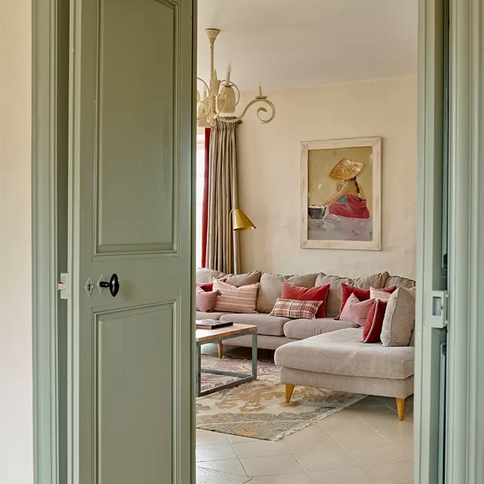 A cosy living room inside La Roussie Villa, viewed through open double doors. It features a beige sectional sofa with red and plaid cushions, a wooden coffee table, and an abstract painting on the wall. The room has a soft, neutral colour palette and a decorative chandelier.