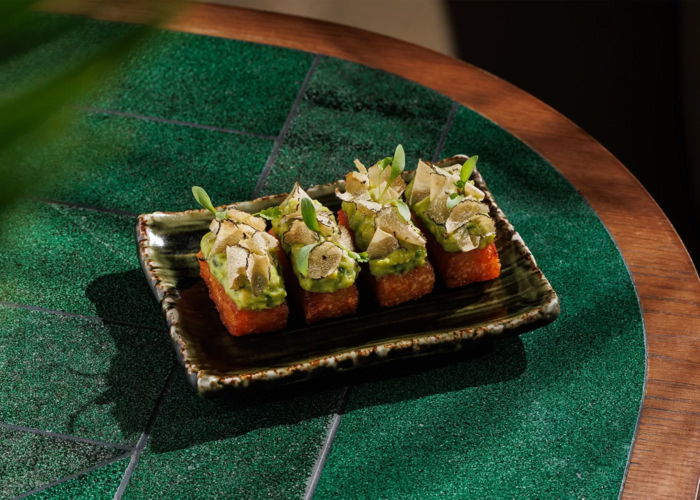 Four pieces of crispy rice topped with avocado, thinly sliced mushrooms, and microgreens are arranged on a rectangular ceramic plate, set on a green patterned table.