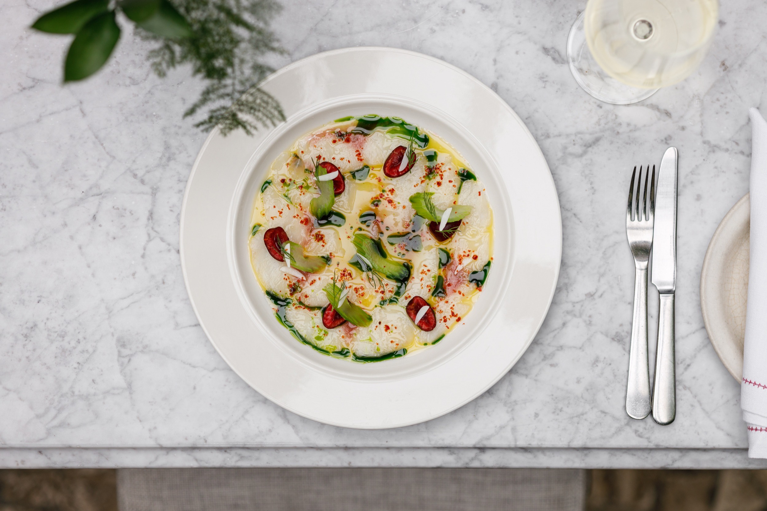 A white plate on a marble table holds a colorful dish of thinly sliced fish, garnished with green herbs and red chili slices. A glass of white wine, a fork, and a knife are placed nearby. A small plant is partially visible in the top left corner at Luca, Farringdon.