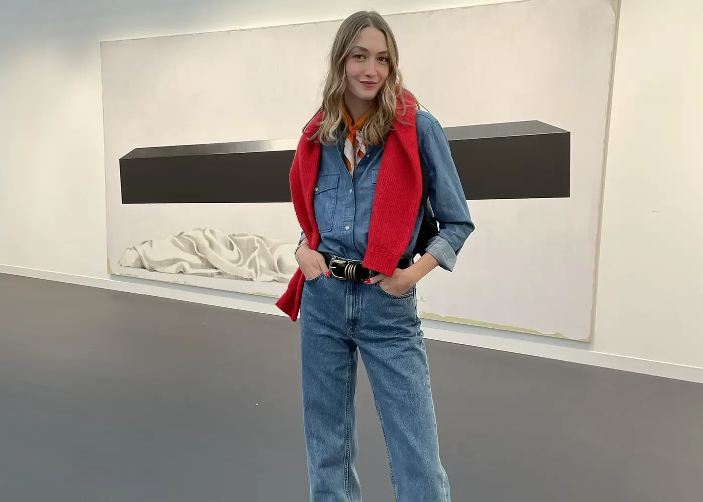 A woman stands indoors wearing a denim shirt and jeans, with a red sweater draped over her shoulders. She poses with one hand in her pocket, smiling slightly. Behind her is a large, minimalist artwork on the wall.