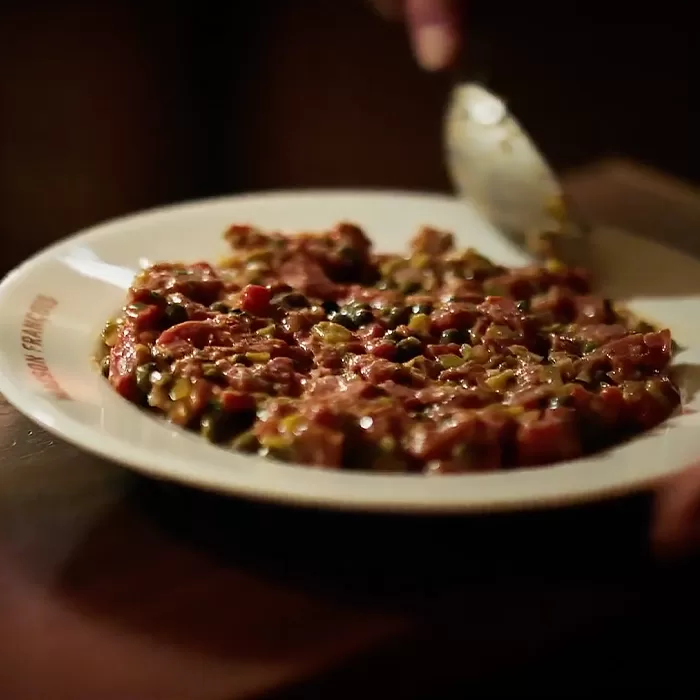 A close-up of a plate with steak tartare, topped with capers and seasonings, as a spoon is held above the dish, ready to serve.