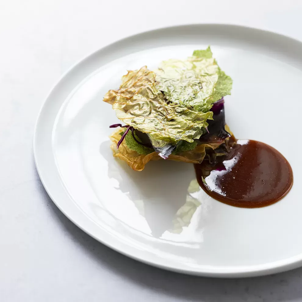 A gourmet dish featuring delicately arranged leafy greens atop a small portion of sauce on a white plate. The presentation is minimalist and elegant, with the sauce artfully placed beside the greens.