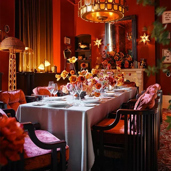 A warmly lit dining room features a long table set with glassware and an arrangement of lush, orange flowers. The room is decorated in rich reds and browns, with plush chairs, and a chandelier casting a soft glow, creating an inviting ambiance at Midland Grand: Private Dining Room, London.