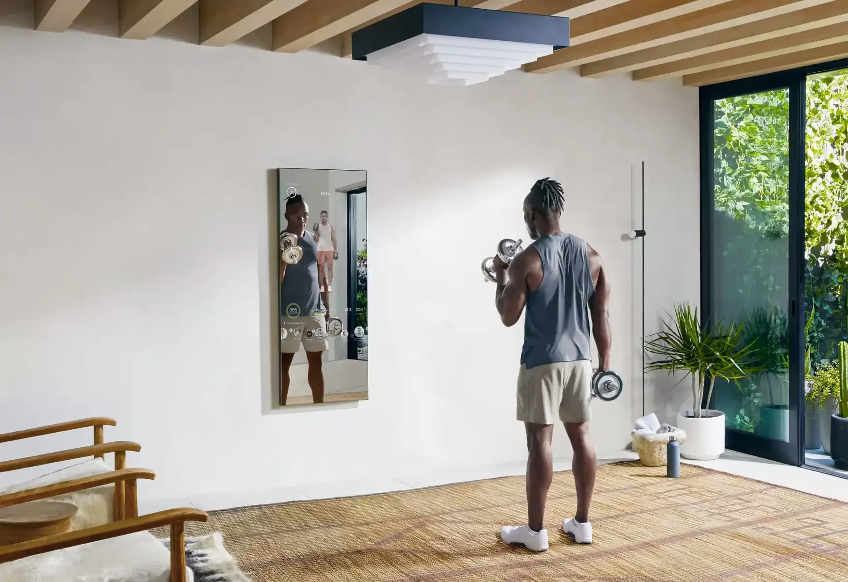 Man in gym clothes with weights working out opposite a home fitness technology mirror in a sunny room