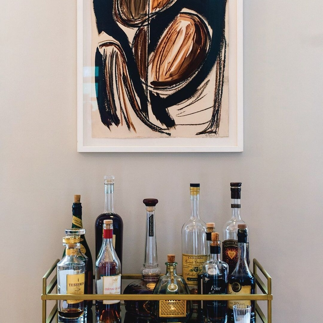 A stylish bar cart with various bottles of liquor and a glass sits against a wall. Above it hangs an abstract painting with bold brown and black brushstrokes. The cart features a metallic frame and glass shelves at The Clove Club, Shoreditch.
