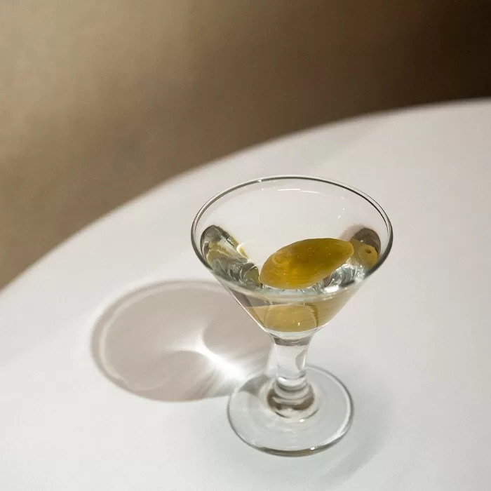 A clear martini glass with a green olive inside sits on a white surface, casting a shadow to the left.