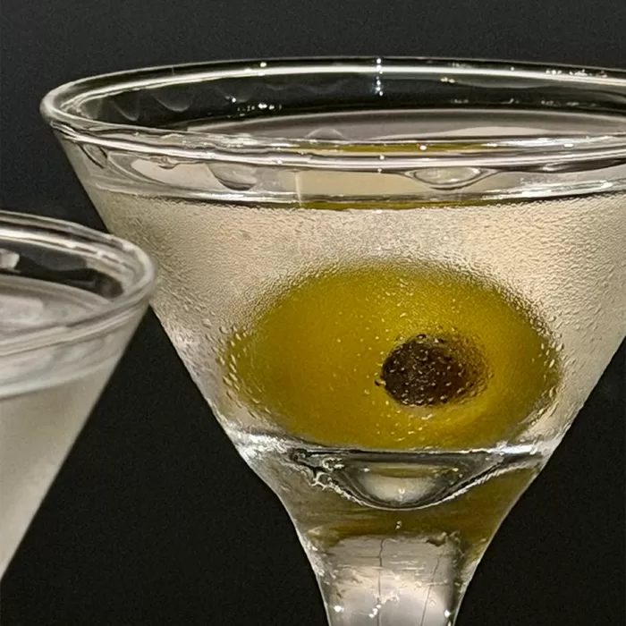 A close-up of a martini glass filled with a clear drink and garnished with a green olive, set against a dark background. Another partially visible martini glass is on the left side.