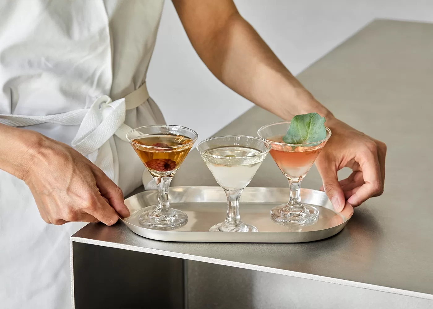 A person in a white apron holds a silver tray with three small martini glasses, each containing a different colored cocktail; one is amber, one is clear, and one is pink with a green leaf garnish.