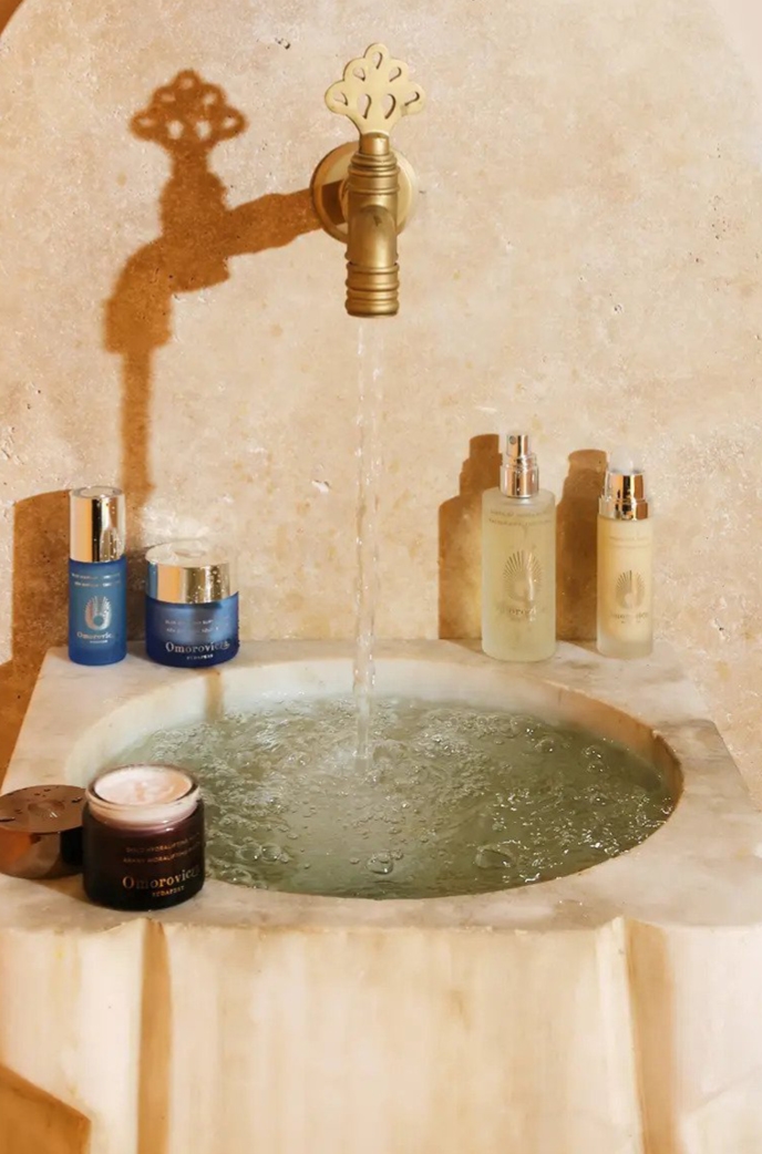 Granite circular sink filled with water with a golden tap and beauty products surrounding at Omorocviza, Budapest