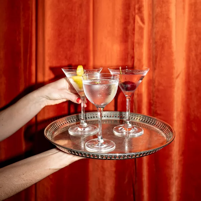 A person holds a round silver tray with three martini glasses, each filled with clear or slightly tinted cocktails and garnished with lemon twist, olive, and cherry, in front of an orange curtain.