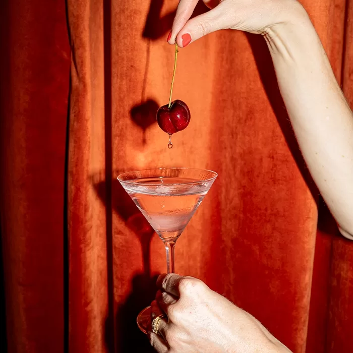 A hand holds a martini glass with a clear drink, while another hand dangles a red cherry above it, with a drop about to fall. The background is an orange-red curtain.