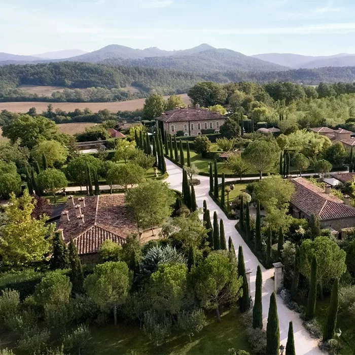 Aerial view of a lush estate with tree-lined pathways, Mediterranean-style buildings, and cypress trees, surrounded by fields and forested hills under a clear sky.