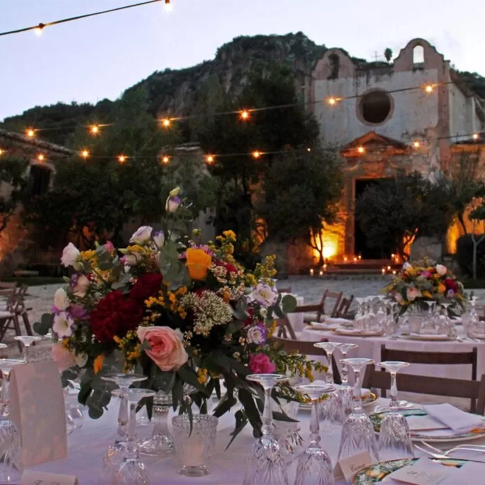Outdoor evening wedding reception with round tables set with floral centerpieces, glasses, and plates. String lights hang above, and a rustic building with arched windows is in the background, surrounded by trees.