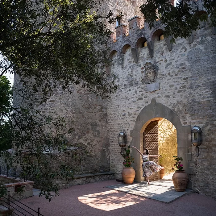 A woman in a flowing dress stands by the arched entrance of a stone castle, surrounded by large potted plants and dappled sunlight. Trees frame the scene, adding to the serene and historic atmosphere.