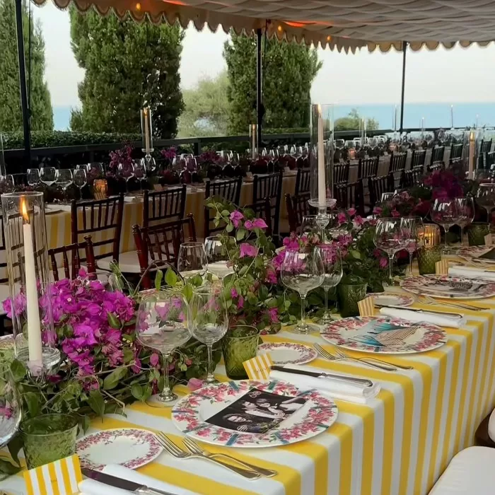 A long, elegantly set outdoor table features yellow and white striped tablecloths, floral plates, lush purple flower centerpieces, glassware, and tall candles, with greenery and a body of water visible in the background.