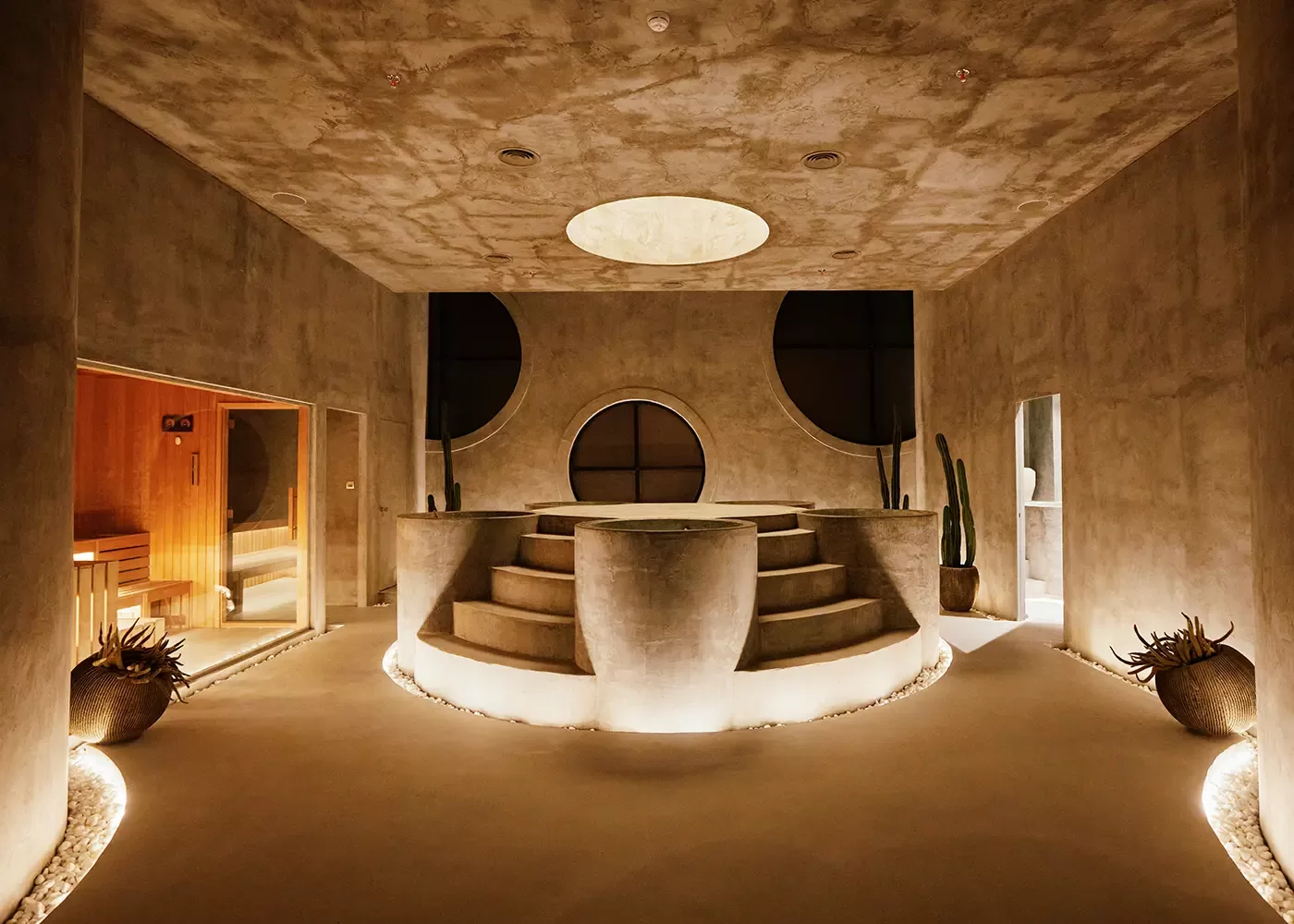 A modern, minimalist spa room with concrete walls and floors, circular windows, a large raised round tub in the center with steps, soft lighting, potted cacti, and dry plants along the sides.