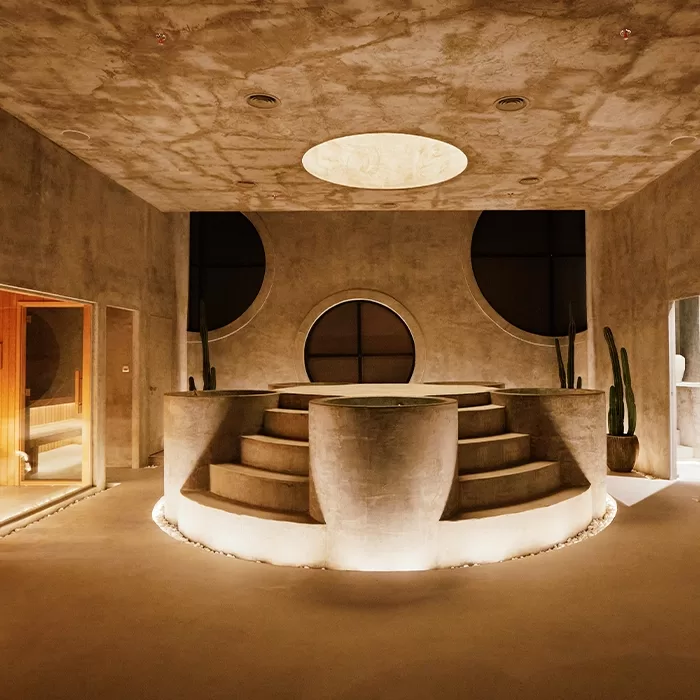 A modern, minimalist spa room with a large circular hot tub in the center, surrounded by curved steps, soft lighting, circular wall cutouts, and potted cacti. The walls and ceiling are finished in textured concrete.