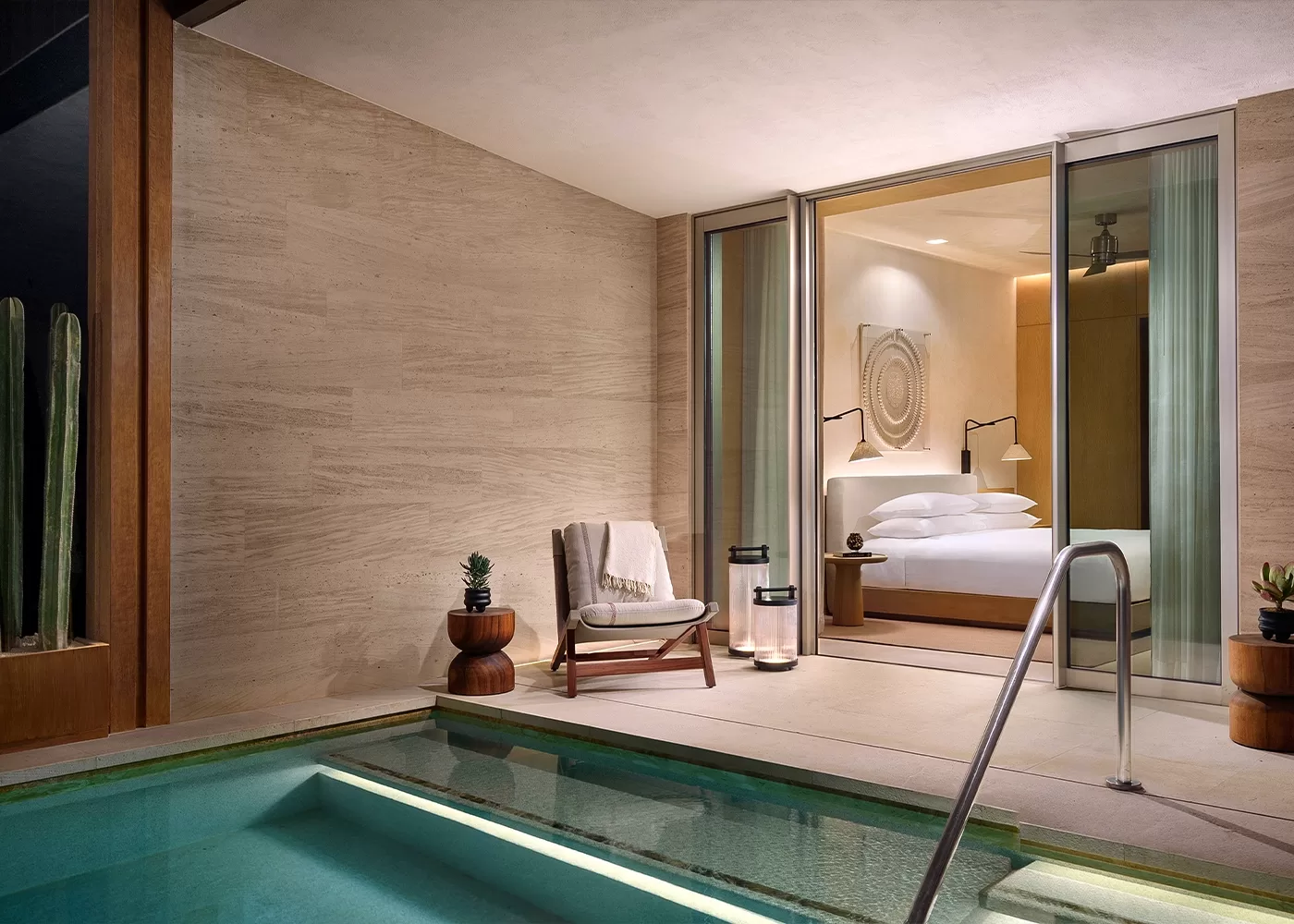 A modern hotel room with sliding glass doors opens to a private patio featuring a chair, small tables with plants, and a pool with a metal handrail. The room has neutral tones and contemporary decor.