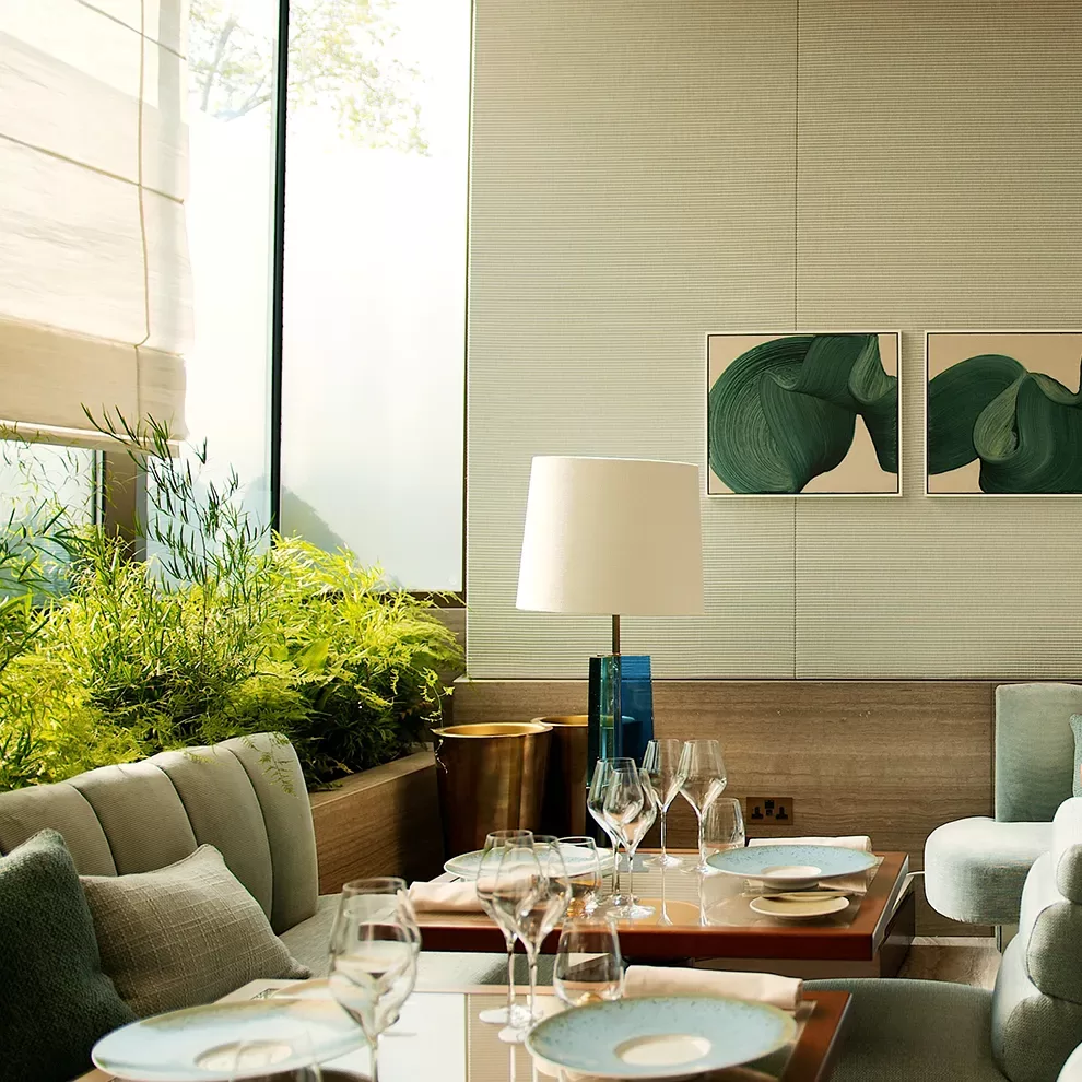 A cozy restaurant interior with a modern design. A table is set with white plates, wine glasses, and cutlery. Soft green plants line the window, and abstract green art pieces hang on a light-colored wall. Natural light fills the space.