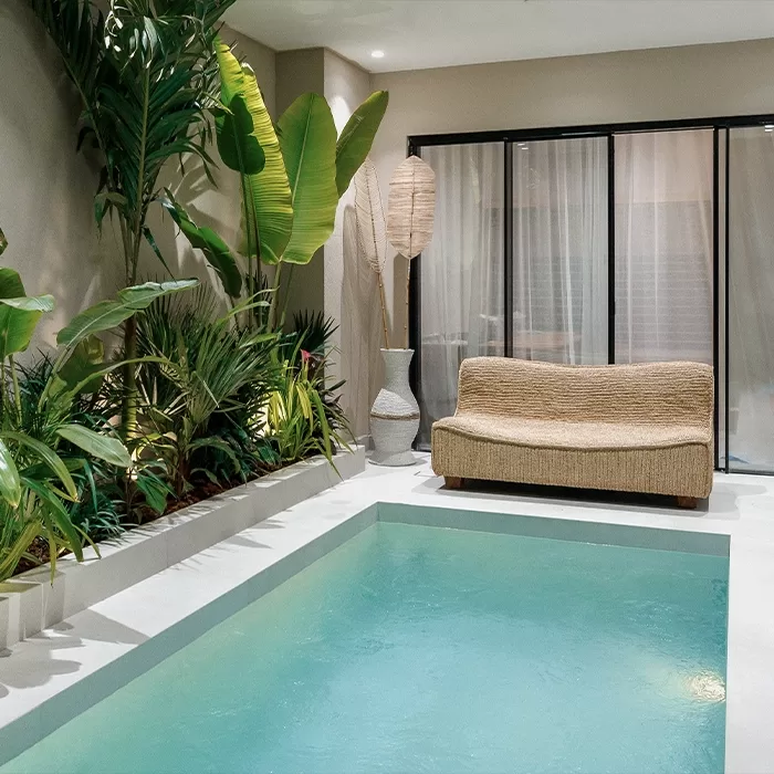 A small indoor pool surrounded by tropical plants, with a woven sofa and a tall decorative lamp placed near floor-to-ceiling glass doors with sheer curtains. The space has a relaxing, modern atmosphere.