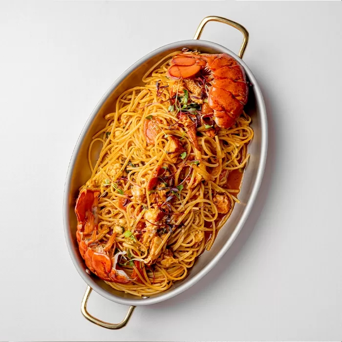 Oval dish with spaghetti topped with lobster in a red sauce, garnished with herbs, on a white background.