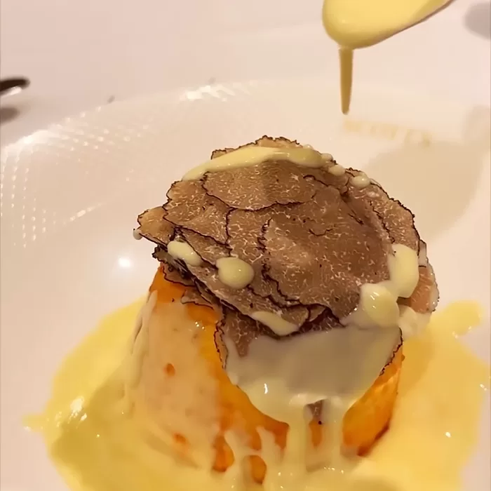 A gourmet dish topped with thin truffle slices is being drizzled with creamy yellow sauce on a white plate.