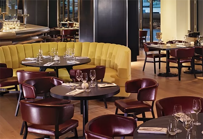 Elegant restaurant interior with a mix of yellow booth seating and dark wooden chairs around round tables. Place settings include wine glasses and cloth napkins. Large windows in the background offer a view outside at Sea Containers, London.