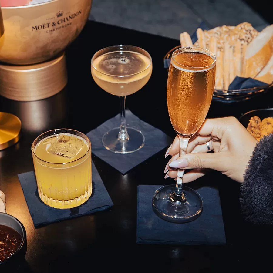 Three different cocktails on a black table with a hand holding a champagne flute, surrounded by snacks, breadsticks, and a Moët & Chandon champagne bucket in the background.