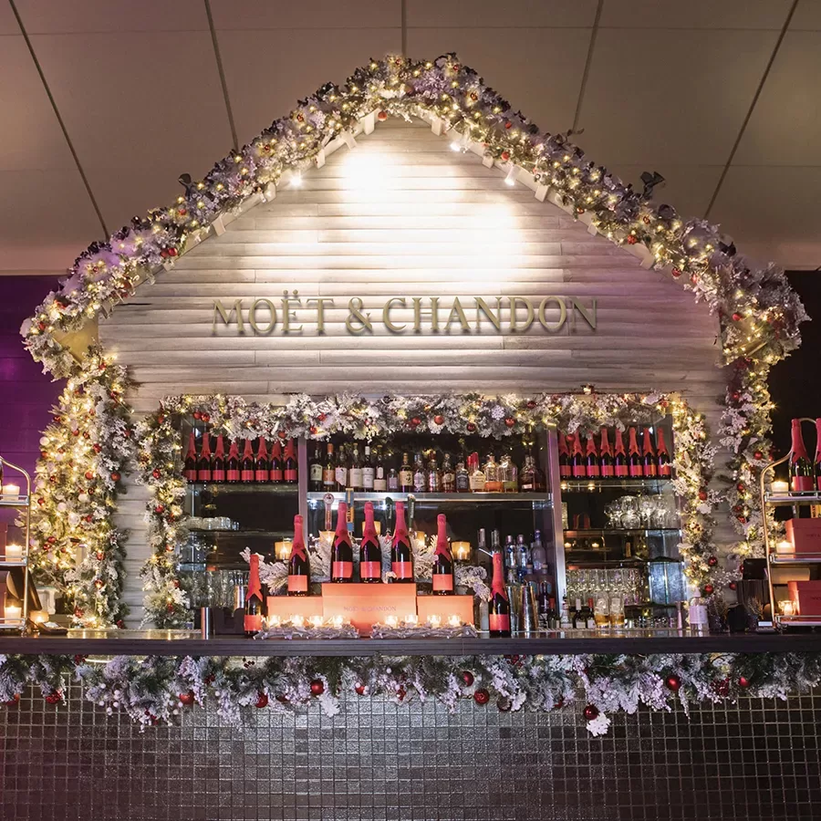A festive bar decorated with garlands, lights, and ornaments, featuring Moët & Chandon branding. Bottles of Moët champagne are displayed on shelves behind the bar, creating a holiday atmosphere.