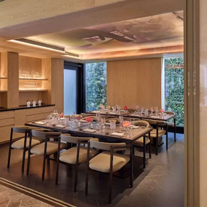 A modern dining room with a long rectangular table set for a meal, featuring neatly arranged glassware and plates. Wooden chairs surround the table, and the room has soft lighting and large windows with views of greenery outside at Akira Back at Mandarin Oriental Mayfair, London.