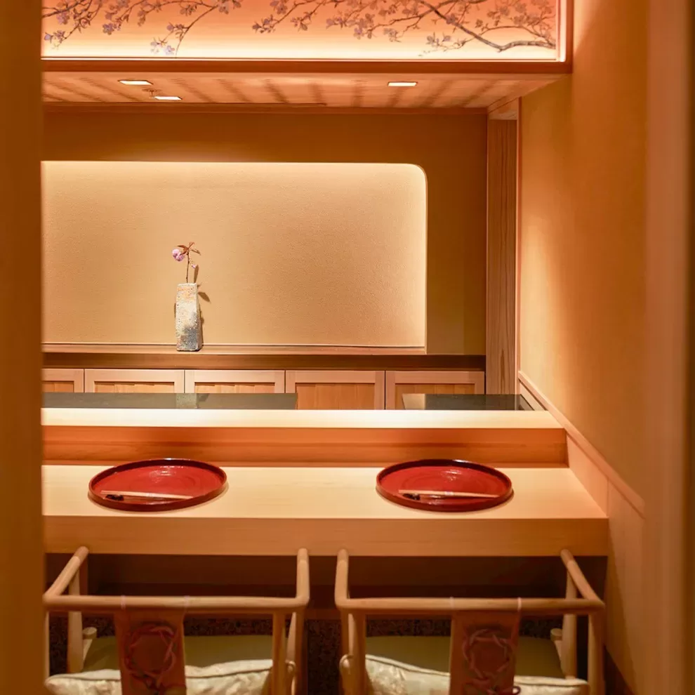 A cozy Japanese restaurant interior with two wooden chairs at a bar counter, featuring red plates. A decorative vase with a single purple flower sits on a recessed shelf, and a cherry blossom design is on the lighted ceiling panel.