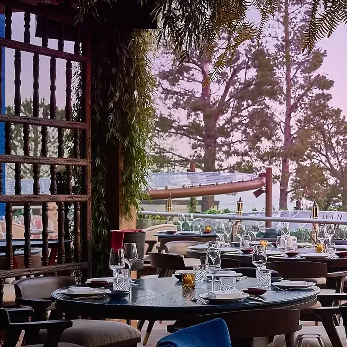 An outdoor dining area with a stunning view of the ocean at sunset. The setting is casual yet elegant, with round tables surrounded by cushioned chairs. The tables are arranged on a terrace overlooking the water, with trees and greenery framing the scene. The sky is painted in soft pastel hues as the sun sets, creating a serene and picturesque atmosphere at COYA Monte-Carlo.