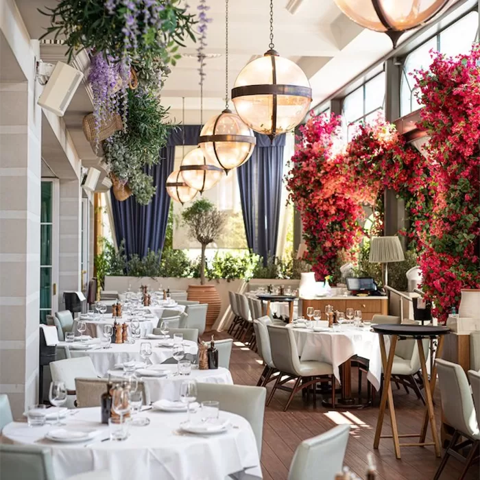 A bright and airy indoor restaurant space adorned with lush greenery and vibrant flowers. The ceiling features large, spherical light fixtures, while the walls are lined with large windows letting in plenty of natural light. The dining area is filled with neatly arranged tables covered in white tablecloths, surrounded by comfortable white chairs. The decor is fresh and inviting, with an emphasis on natural elements, including hanging plants and floral arrangements at GAIA Monte-Carlo.